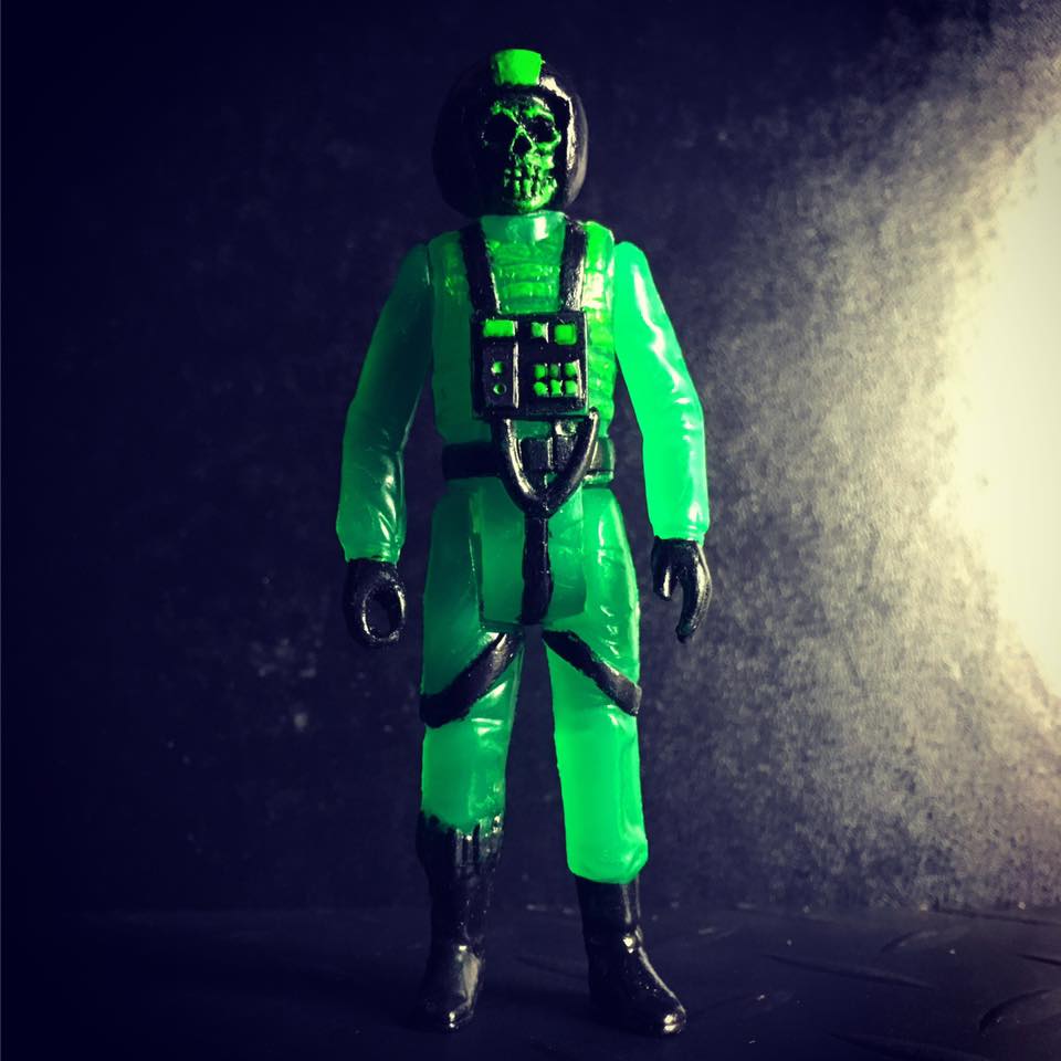 Small glow-in-the-dark green plastic figure of a humanoid soldier with a skull face, dark eerie background, moody lighting